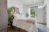 Bright and airy bedroom with modern decor, featuring a cozy double bed, chic abstract artwork, and lush green plants, enhanced by natural sunlight through large windows.