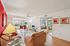 Bright, cozy living room with modern decor, featuring a red accent wall, glass-top tables, and plush seating. Abundant natural light flows in from large windows, leading to a glimpse of a stylish bedroom.