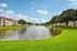 Serene apartment complex with picturesque pond, lush greenery, and elegant multi-story buildings under a clear blue sky.