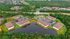 Aerial view of a serene apartment complex with modern buildings nestled around a picturesque pond, surrounded by lush greenery and ample parking space, offering a tranquil and inviting living environment.