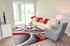 Modern living room with a chic white couch, vibrant red and gray accents, glass coffee table, and a stylish area rug, overlooking a sunlit patio.