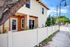 Charming two-story home with a warm peach and cream facade, nestled behind a pristine white fence, surrounded by lush greenery and shaded by a large tree, creating an inviting and serene street view.