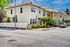Charming corner townhouse with a sunny facade, surrounded by lush greenery and a welcoming white fence, perfect for rental opportunities.