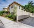 Charming two-story yellow building surrounded by lush greenery and palm trees, featuring a modern white fence for privacy in a sunny neighborhood.