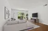Bright and airy living room with modern decor, featuring a cozy L-shaped sofa, elegant wall art, and a large window offering a lush outdoor view.
