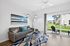 Bright and modern living room with sleek glass coffee table, cozy gray sofa, and vibrant artwork, opening to a lush green outdoor space through sliding glass doors.