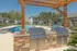 Charming outdoor space featuring a sparkling pool with sun loungers, a stone BBQ area, and lush landscaping under a clear blue sky.