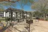 Charming outdoor community area with shaded pergola, picnic tables, and built-in grills set against a backdrop of lush greenery.