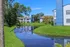 Serene apartment complex with charming blue and white buildings, nestled by a picturesque pond and lush greenery under a clear blue sky.