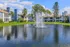 Charming lakefront apartment complex with lush greenery and a sparkling fountain under clear blue skies.