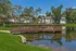 Scenic apartment complex with charming wooden bridge overlooking a tranquil pond, surrounded by lush greenery and modern homes.