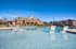 Luxurious apartment complex with a sparkling resort-style pool featuring modern lounge chairs, surrounded by beautiful brick architecture and clear blue skies.