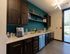 Modern kitchenette with sleek dark cabinetry, vibrant teal accent wall, and ample natural light from large window.