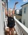Joyful scene of a young woman with a backpack celebrating on a sunlit balcony of a modern apartment, surrounded by stylish buildings and lush greenery.