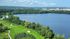 Scenic view of a lush green golf course beside a tranquil lake, surrounded by verdant forests and a distant city skyline under a blue sky.