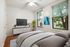 Inviting bedroom with a stylish decor, featuring large windows offering natural light, a cozy bed with plush bedding, a modern TV setup, and elegant artwork complementing the serene ambiance.