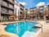 Luxurious apartment complex with a sparkling pool and sun loungers, surrounded by modern multi-story buildings and vibrant orange umbrellas under a clear blue sky.