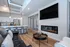 Modern apartment interior featuring a sleek living room with a black leather sofa, wall-mounted TV, and stylish electric fireplace. Open kitchen with contemporary bar stools and stainless steel appliances, enhanced by bright lighting and wooden flooring.