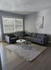 Elegant living room with modern gray sofa, matching armchair, and sleek coffee tables on a plush rug, bathed in natural light from large windows.