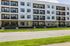 Modern apartment building with large windows and private balconies, surrounded by lush greenery and a pristine sidewalk.