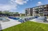 Modern apartment building with stylish pool area featuring blue lounge chairs, umbrellas, and vibrant green landscaping under a bright blue sky.