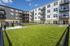 Bright and modern apartment complex with beautifully landscaped green area, sleek balconies, and inviting outdoor seating, set against a clear blue sky.