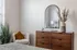 Elegant bedroom corner with chic wooden dresser, arched mirror, cozy chair, and decorative plants creating a serene ambiance.