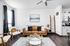 Modern living room with a cozy tan sofa, elegant glass coffee table, and stylish decor, complemented by a sleek kitchen backdrop.