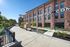 Charming red brick building with large windows, set in a vibrant urban environment featuring a landscaped walkway and park benches, ideal for modern urban living.