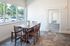 Bright dining area with rustic wooden table, large windows offering lush garden views, and modern tiled floor.