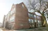 Charming red brick apartment building with large windows and an inviting entrance, surrounded by mature trees and a sunny pathway.