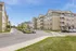 Modern apartment complex with charming beige and gray exteriors, featuring private balconies, landscaped surroundings, and ample parking, set against a clear blue sky.