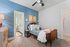 Charming bedroom with vibrant blue accent wall, cozy twin bed adorned with plush toys, and a view into a brightly lit bathroom with floral wallpaper.