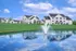 Picturesque apartment complex with modern buildings and a serene pond featuring a vibrant fountain, set against a clear blue sky.