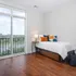 Bright bedroom with sleek wood flooring, featuring a cozy bed with colorful pillows, modern side lamp, and a scenic view of lush greenery through floor-to-ceiling windows.