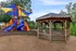 Charming outdoor area featuring a vibrant children's playground with colorful slides and climbing structures, next to a picturesque wooden gazebo under a clear blue sky.