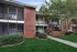 Charming apartment exterior featuring red brick accents, lush greenery, and a welcoming pathway under a sunny sky.