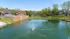 Tranquil housing community featuring a serene pond with a decorative fountain, lush greenery, and cozy brick buildings in the background.