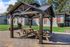 Charming outdoor space with a wooden gazebo and picnic tables, surrounded by lush greenery and modern apartments.