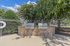 Outdoor barbecue area with two sleek stainless steel grills set against a stone backdrop, surrounded by lush greenery and a gated fence.