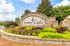 Charming stone entrance of Summer Lake Luxury Apartment Homes surrounded by lush greenery and vibrant flowers under a bright blue sky.