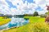 Stunning view of serene lakeside apartments with lush green surroundings and picturesque reflections under a vibrant blue sky.