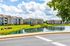 Charming apartment complex with modern architecture, nestled by a tranquil pond and lush green landscape, under a bright blue sky.