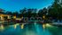 Luxurious apartment complex pool area at dusk with glowing lights, surrounded by lush greenery and elegant cabanas.