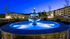 Serene twilight scene featuring an illuminated tiered fountain set against modern apartment buildings with lush landscape.