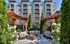 Elegant outdoor seating area with cozy sofas around a stylish fire pit, surrounded by lush greenery and modern apartment building in the background.