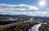 Breathtaking aerial view of a scenic cityscape with lush greenery, a shimmering river, and mountain backdrop under a bright, sunny sky.