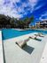 Inviting swimming pool area with elegant lounge chairs, surrounded by lush greenery and modern apartment buildings, under a vibrant blue sky.