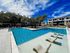 Luxurious apartment complex with a sparkling swimming pool, surrounded by lush trees and modern multi-story buildings under a clear blue sky.