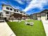 Charming apartment complex with modern architecture, featuring a lush green courtyard, shaded seating area, and vibrant blue sky.